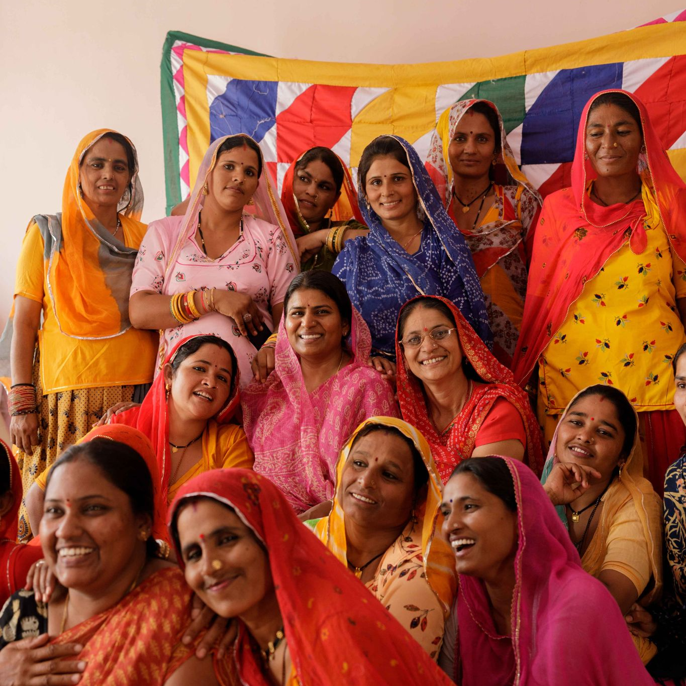 Exploitative labor practices: Image of a large group of artisan women standing outside of an Indian garment workplace. They are all smiling and happy, and some are holding hands or touching their shoulders.